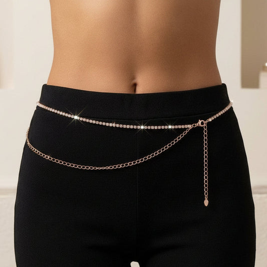 A close-up of a model's lower torso wearing a delicate rose gold chain waist belt over black leggings, with a softly blurred indoor background.